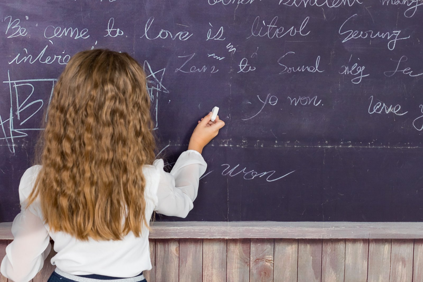 writing on blackboard. girl in class.school desk, education concept.hand holding a chalk on the chalkboard.back view of a little girl writing with chalk on the backboard in school class